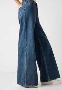 Jeans en denim à jambes larges en bleu foncé. Comprend une taille haute, de grandes poches et un ourlet effiloché. Le matériau a une légère texture avec des coutures visibles.