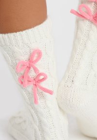 Los calcetines blancos de punto cableado presentan dos lazos de punto rosas a los lados, texturizados para proporcionar calidez y comodidad, con puños acanalados.