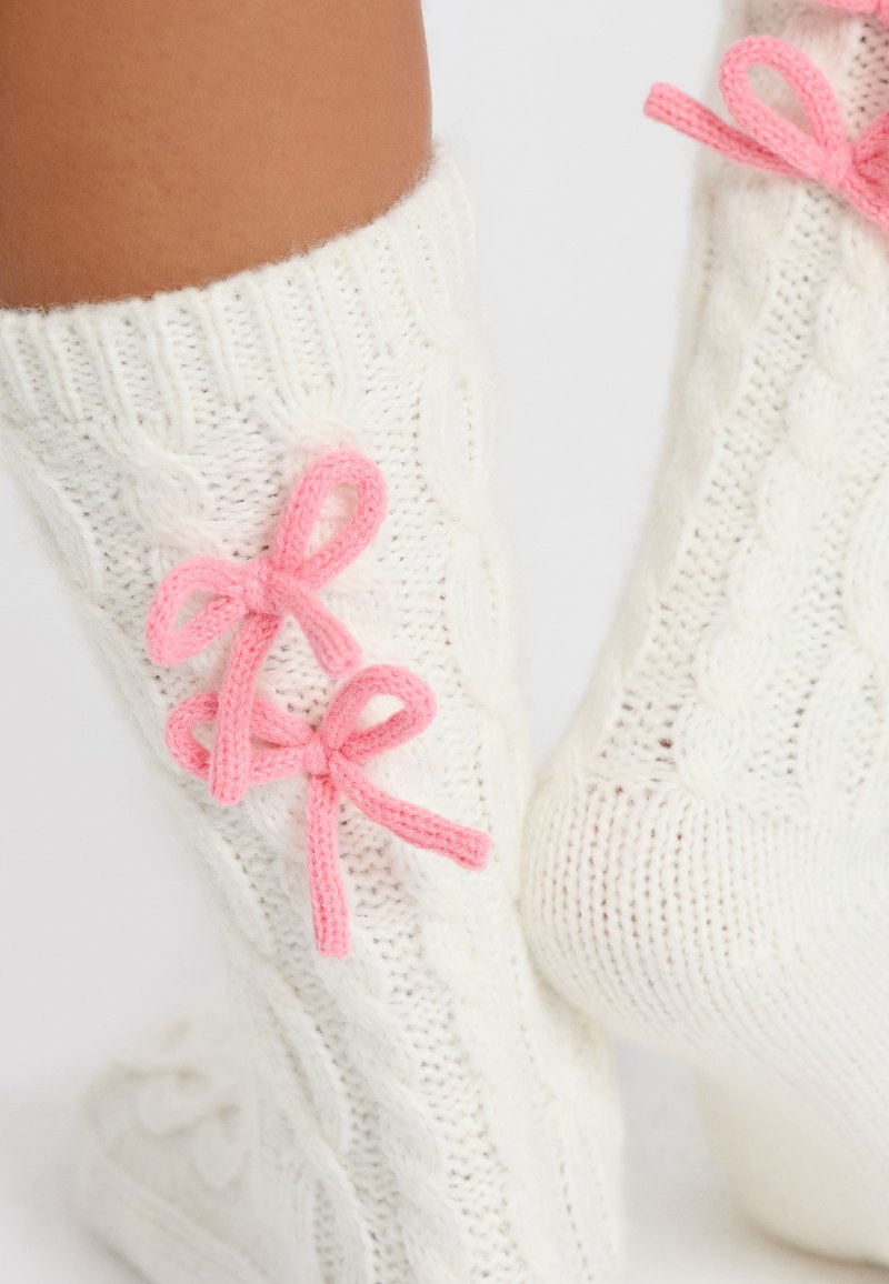 Los calcetines blancos de punto cableado presentan dos lazos de punto rosas a los lados, texturizados para proporcionar calidez y comodidad, con puños acanalados.