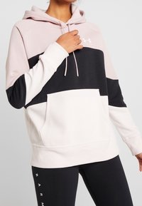 Sudadera con bloques de color en rosa suave, negro y gris claro. Presenta un bolsillo tipo canguro en el frente, capucha con cordón y puños acanalados. Textura de tela suave.