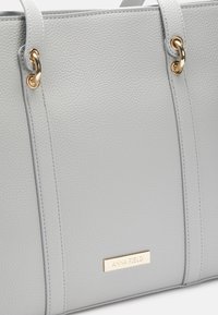 Šedá kabelka s texturou, zdobená zlatými kovovými doplňky, obsahující logo značky a prošité linie pro zvýraznění detailů.