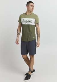 Maslinasto zelena majica s bijelim grafičkim natpisom, u kombinaciji s tamnoplavim kratkim hlačama. Model nosi crne tenisice. Tkanina izgleda mekana, s opuštenim krojem.