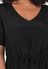 Svart klänning med V-ringning, korta ärmar och knytning i midjan. Materialet verkar mjukt med en lätt glans och ett flödande textur.