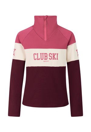 Jong meisje draagt een paarse en roze skipullover met de tekst "Club Ski" en zwarte skipakken met witte sterrenpatronen, staand tegen een effen achtergrond.