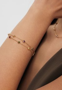 Złota bransoletka z wieloma kolorowymi kamieniami szlachetnymi (fioletowym, różowym, żółtym) na delikatnym łańcuszku, noszona na skórze w odcieniu ciała o gładkiej teksturze.
