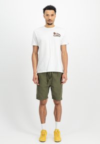Hvid bomulds T-shirt med broderet design, parret med olivengrønne shorts. Modellen bærer gule sneakers og hvide sokker.