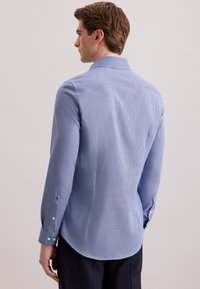 Man in een nauwsluitend lichtblauw gestructureerd overhemd met lange mouwen, gezien vanaf de achterkant tegen een effen lichte achtergrond.
