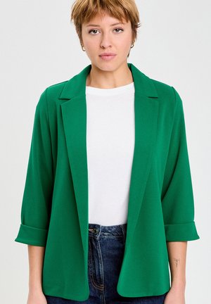 Kobieta o krótkich włosach, ubrana w zieloną marynarkę na białą koszulę i niebieskie dżinsy, stojąca na jednolitym jasnym tle.