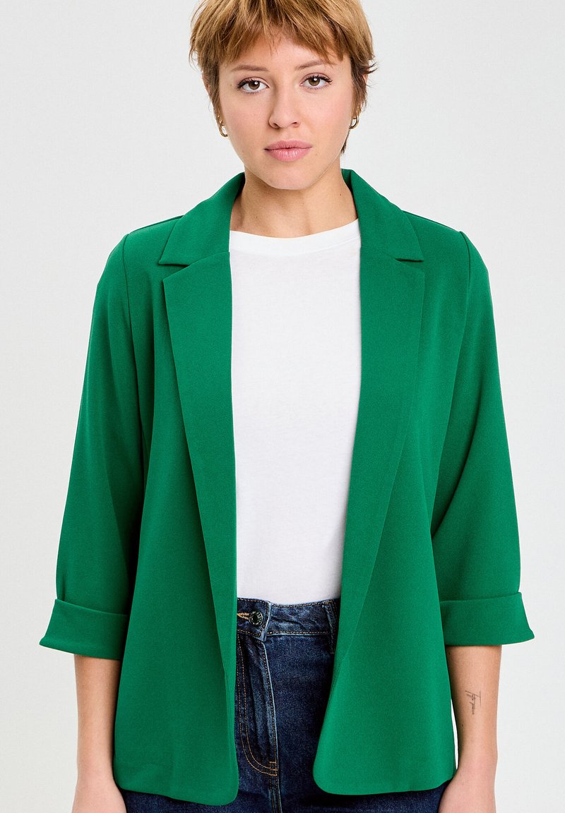 Kobieta o krótkich włosach, ubrana w zieloną marynarkę na białą koszulę i niebieskie dżinsy, stojąca na jednolitym jasnym tle.