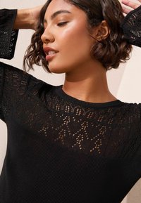 Juoda mezgimo palaidinė su tekstūruota nėrinių dizainu ant pečių ir viršutinės krūtinės, su raukšlėtomis rankovėmis ir iškirpte.