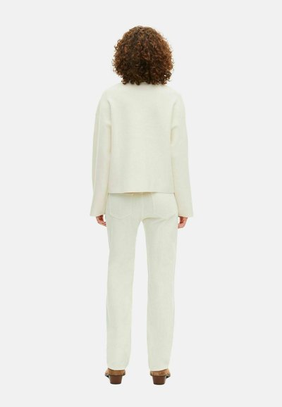 Donna con capelli ricci castani che indossa un maglione a maniche lunghe bianco sporco, pantaloni abbinati e scarpe basse marroni, in piedi, voltata di spalle.
