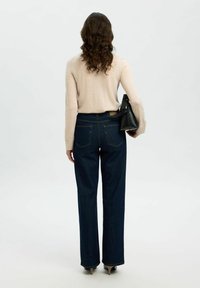 Jeans wide-leg in denim blu scuro con cuciture gialle a contrasto, abbinati a un top beige a maniche lunghe, completati con una borsa nera.