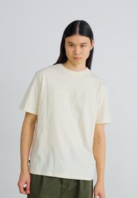 Ljust beige bomullströja med "Timberland"-logotypen i en något upphöjd, tonad tryck. Korta ärmar och klassisk rundad halsringning.