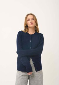 Navyblauwe gebreide cardigan met een ronde hals, knoopsluiting aan de voorkant, ribgebreide manchetten en twee voorzakken. Zachte textuur en getailleerd ontwerp.