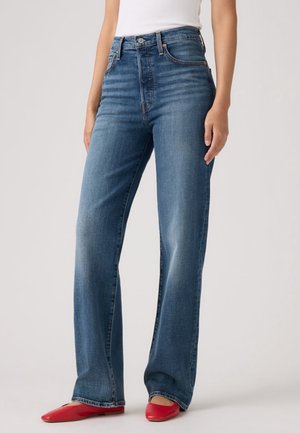 Femme portant un jean bleu taille haute à jambes droites et des chaussures plates rouges pointues sur un fond uni.