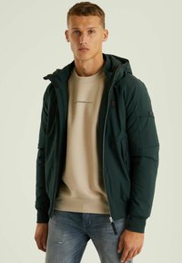 Temno zelena jakna s kapuco in zadrgo, stranskimi žepi in elastičnimi manšetami, nošena čez bež sweatshirt in svetlo modre kavbojke.