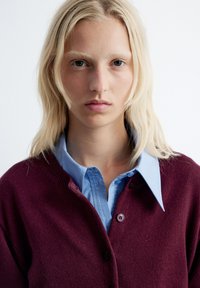 Bordeauxfarbener Cardigan über einem hellblauen Hemd, mit Knopfverschluss und gerippten Details am Kragen. Glatte Textur, schlichtes Design.