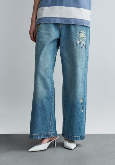 Jeans blu a gamba larga in denim, con dettagli ricamati di un sole e una montagna, con leggeri effetti usurati e orlo sfrangiato.