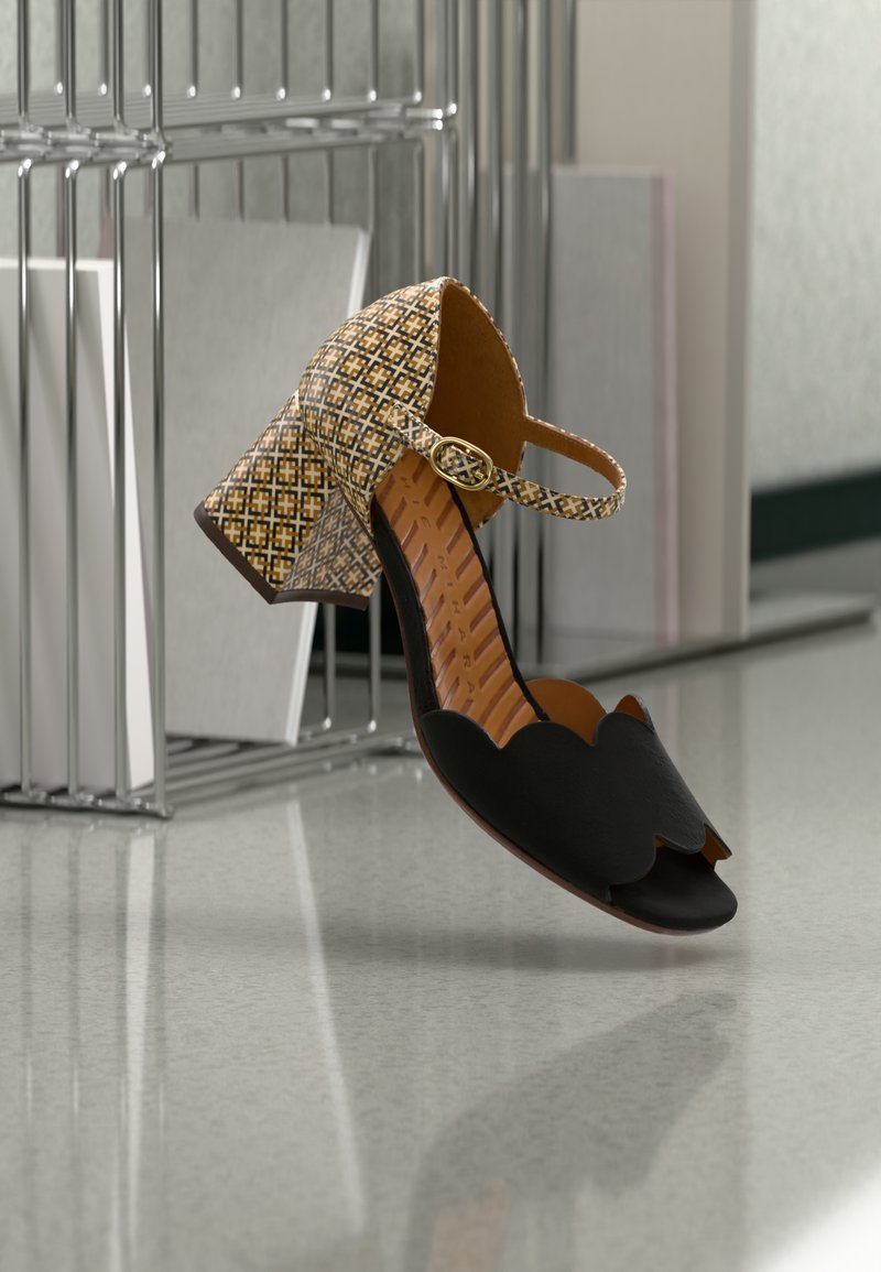 Chaussure noire à bout ouvert avec bords festonnés, présentant un talon et une bride de cheville beige à motif avec une boucle en or. Finition en cuir lisse.