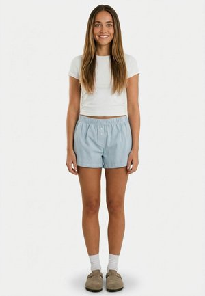 Ung kvinde med langt brunt hår iført hvid crop top, lyseblå shorts, hvide strømper og beige slip-on sko, stående og smilende.
