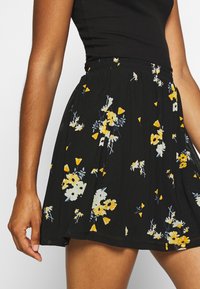 Zwarte bloemenrok met een geplooide taille, versierd met gele en witte bloemen op een textuurstof. Elastische tailleband voor comfort.