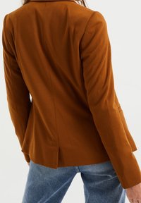 Brown fitted blazer with a structured design, featuring a single vent at the back and soft fabric. Paired with light blue denim jeans.