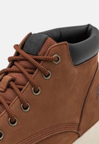 Botín de cuero marrón con un acabado texturizado, que cuenta con cordones marrones, ojales de metal y un cuello acolchado negro. Logo de Timberland en el lateral.