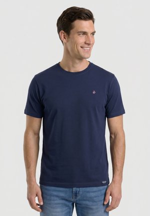 Hombre sonriendo y mirando hacia la derecha, con una camiseta lisa de manga corta azul marino y vaqueros azul claro sobre un fondo liso.