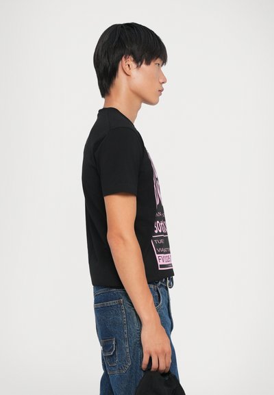 T-shirt noir avec texte graphique rose sur le devant, manches courtes et encolure ras du cou, associée à un jean en denim bleu.