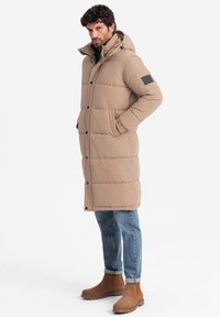 Abrigo largo acolchado beige con capucha, bolsillos laterales y un parche con el logo en la manga, combinado con jeans azules y botas de tobillo marrones.