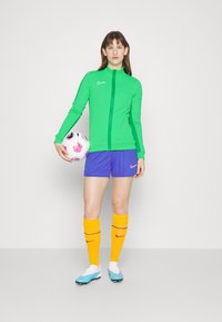 Grüne Zip-Jacke, blaue Shorts, gelbe Socken und Fußballschuhe. Hält einen mehrfarbigen Fußball. Nike-Logos auf der Kleidung.