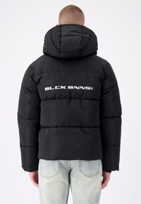 Zwarte gewatteerde jas met capuchon, voorzien van gestikte secties en een wit logo op de achterkant. Draag met lichtblauwe jeans.