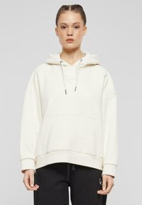 Cream-colored hoodie made of soft fabric, featuring a front pocket, drawstring hood, and subtle logo embroidery on the chest.