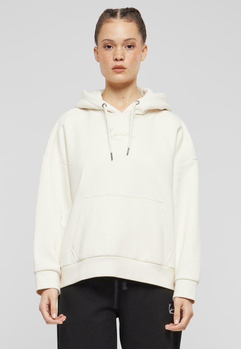 Cream-colored hoodie made of soft fabric, featuring a front pocket, drawstring hood, and subtle logo embroidery on the chest.