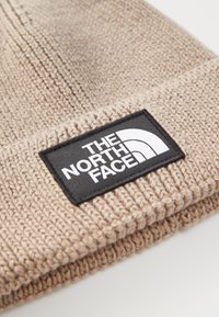 Beige kudumisega beanie, mille ääres on volditud cuff ja must ristkülikukujuline silt valge "The North Face" logoga.