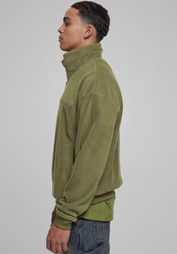 Giacca in pile verde oliva con colletto alto, zip frontale, tasche laterali e polsini a costine, indossata su una maglietta verde.