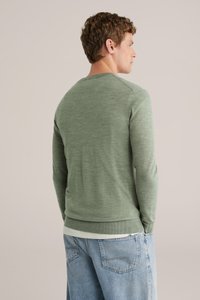 Grön långärmad tröja med ribbad halsringning och muddar, kombinerad med ljusblå jeans, visad bakifrån. Mjuk textur.