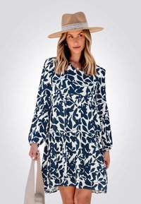 Robe longue à manches longues florale bleu marine et blanche avec une jupe à volants et un corsage froncé, associée à un chapeau à larges bords beige et un sac à main neutre.