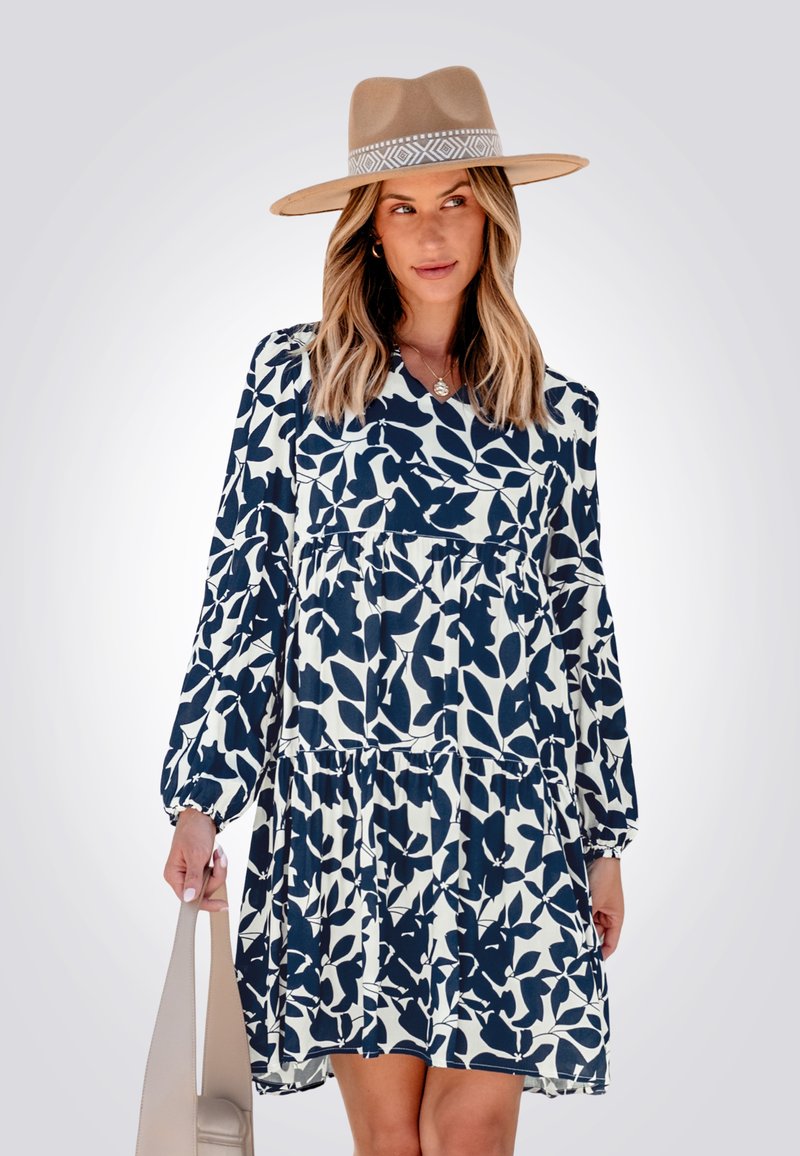 Abito lungo a maniche lunghe navy e bianco con una gonna a strati e corpetto arricciato, abbinato a un cappello a tesa larga color sabbia e una borsa neutra.
