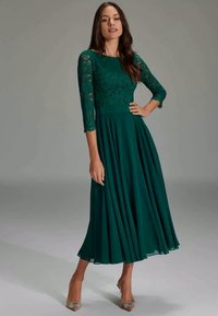 Robe longue verte avec un corsage en dentelle et des manches trois quarts, jupe fluide et plis. Portée avec des chaussures à talons beiges.