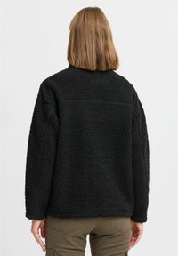 Black fleece jacket with a textured surface, relaxed fit, high collar, and drop shoulders, paired with olive green cargo pants.
