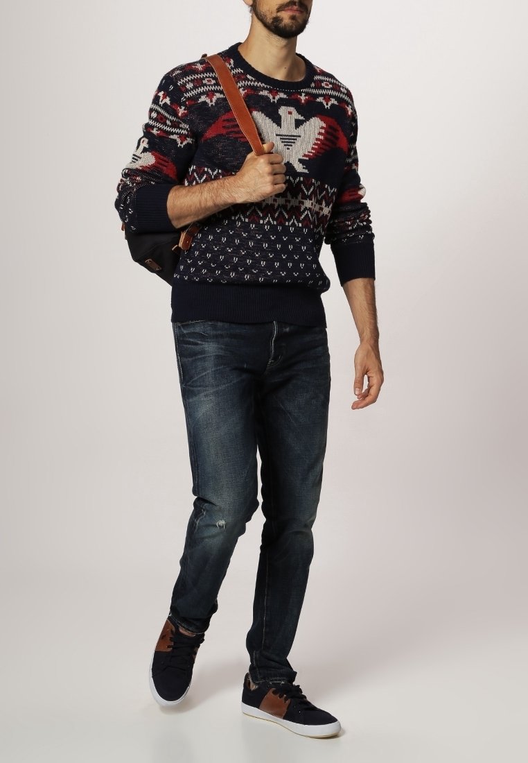 Námorníkově modrý pletený svetr s vícebarevnými vzory, modré džíny s opotřebovanými detaily a tmavé tenisky s hnědými akcenty.