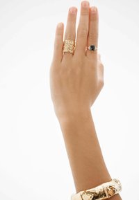 Anillos metálicos en tono dorado con diseños texturizados y de filigrana en una mano, junto a un anillo delgado que presenta una piedra oscura.