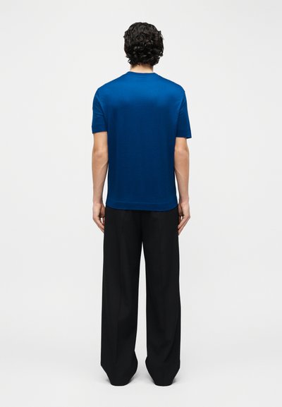 Άνδρας με σγουρά μαύρα μαλλιά, φορώντας μπλε πουκάμισο με κοντά μανίκια και φαρδιά μαύρα παντελόνια, στέκεται με την πλάτη προς το μέρος μας σε απλό λευκό φόντο.