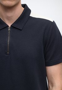 Granatowa koszulka polo z fakturowanej tkaniny, z zamkiem przy kołnierzu i białymi akcentami na ramionach. Krótkie rękawy o nowoczesnym, dopasowanym kroju.