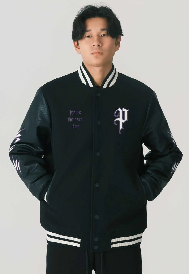 Giacca varsity nera con maniche in pelle, colletto e orlo a coste bianchi a righe, e lettering viola sul davanti. Presenta una grafica sulla manica.