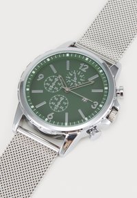 Chronograph-Uhr mit grünem Zifferblatt, silbernem Netzarmband, silbernen Akzenten, großen Stundenmarkierungen und drei Unterzifferblättern für Zeitmessfunktionen.