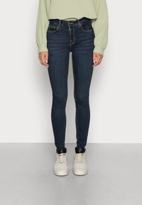 Persona con jeans skinny azul oscuro, camiseta de manga larga verde claro y zapatillas blancas de pie contra un fondo claro y sencillo.