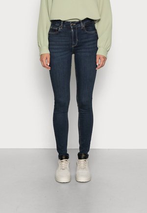 Persoon in donkerblauwe skinny jeans, lichtgroen langemouwen shirt en witte sneakers die tegen een effen lichte achtergrond staat.