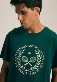 Zielona bawełniana koszulka z centralnym wzorem przedstawiającym krzyżujące się rakiety tenisowe i piłkę tenisową, otoczona wieńcem laurowym oraz tekstem.